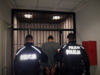 Umundurowani policjanci doprowadzają osobę podejrzaną o udział w pobiciu. Dwóch funkcjonariuszy prowadzi osobę z założonymi z tyłu kajdankami.