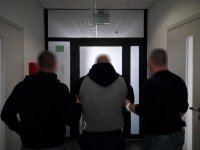 Policjanci doprowadzają osobę podejrzaną o udział w pobiciu. Dwóch funkcjonariuszy prowadzi osobę z założonymi z tyłu kajdankami.