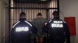 Umundurowani policjanci doprowadzają osobę podejrzaną o udział w pobiciu. Dwóch funkcjonariuszy prowadzi osobę z założonymi z tyłu kajdankami.