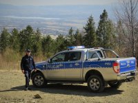 Umundurowany policjant i stoi obok oznakowanego radiowozu Ford Ranger. W tle Beskidy.