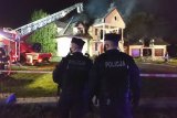 Policjanci zabezpieczają miejsce akcji gaśniczej. W tle płonący budynek i strażacy.