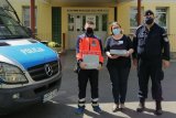 Policjant, ratownik medyczny i pielęgniarka stoją przed szpitalem. Obok widać maskę policyjnego radiowozu.