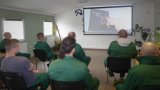 Skazani siedzą w świetlicy zakładu karnego. Na ekranie rzutnika widoczny jest policjant.