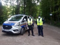 Policjant i Strażnik Miejski stoją przy radiowozie na wjeździe do lasu.