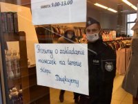 Widok sklepowej witryny z kartką, na której napisana jest prośba o założenie maseczki. Na drugim planie policjant.