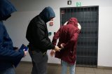 Nieumundurowany policjant zakłada zatrzymanej kobiecie kajdanki na ręce trzymane z tyłu.
