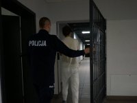 Umundurowany policjant wprowadza zatrzymanego przez otwartą kratę do pomieszczeń