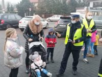 Policjant z ruchu drogowego przekazuję opaskę odblaskową kobiecie pchającą wózek z dzieckiem.