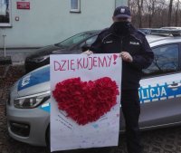 Umundurowany policjant stojący przy radiowozie przed komisariatem trzyma w rekach wyklejankę z kolorowego papieru w kształcie czerwonego serca.