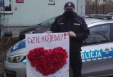 Umundurowany policjant stojący przy radiowozie przed komisariatem trzyma w rekach wyklejankę z kolorowego papieru w kształcie czerwonego serca.