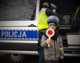 Dziecko trzyma tzw. lizak policyjny, w tle policjant ruchu drogowego i radiowóz.