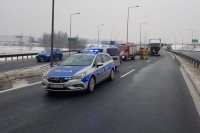 Policyjny radiowóz stoi na drodze przed pojazdem TIR, który brał udział w zdarzeniu drogowym. Na drugim planie pracują policjanci i strażacy.