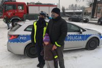Policjantka ruchu drogowego stoi w towarzystwie mężczyzny, który trzyma za ramiona dziecko. W tle radiowóz policyjny.