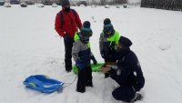 Umundurowany policjant kuca przy dwójce dzieci na śniegu. Zdjęcie wykonane na zboczu stoku narciarskiego.
