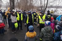 Policjanci i strażacy OSP stoją wśród dzieci na parkingu. Prowadzą pogadankę z gromadką dzieci podczas akcji profilaktycznej.