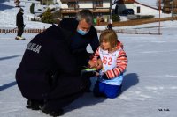 Policjant kuca przy dziecku i rozmawia z nim. Obok kuca opiekun. Zdjęcie wykonane na stoku narciarskim w Beskidach.