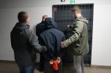 Nieumundurowani policjanci doprowadzają zatrzymanego mężczyznę, stoją przed kratą aresztu otwieraną właśnie przez policjant w mundurze.