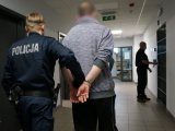 Umundurowany policjant prowadzi zatrzymanego, który ma założone kajdanki na rękach trzymanych z tyłu.