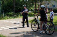 Policjant z ruchu drogowego stoi na starcie trasy egzaminacyjnej na kartę rowerową. Na linii startu stoi dziewczynka z rowerem. Obok niej uczniowie.