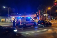 Policjant stoi za radiowozem na sygnałach na drodze i trzyma osobę zatrzymaną. Mężczyzna bez podkoszulka ma kajdanki na rękach z tyłu ciała. Zdjęcie wykonane w nocy.