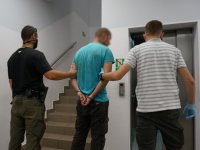 Nieumundurowani policjanci prowadzą zatrzymanego mężczyznę, który ma kajdanki założone na ręce trzymane z tyłu. Stoją przed wejściem do windy.
