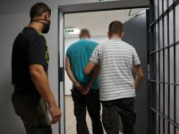 Nieumundurowani policjanci prowadzą zatrzymanego mężczyznę, który ma kajdanki założone na ręce trzymane z tyłu. Wychodzą przez drzwi z kratą na korytarz.
