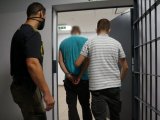 Nieumundurowani policjanci prowadzą zatrzymanego mężczyznę, który ma kajdanki założone na ręce trzymane z tyłu. Wychodzą przez drzwi z kratą na korytarz.
