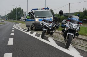 Na pierwszym planie widać dwa stojące na powierzchni wyłączonej z ruchu policyjne motocykle. Na drugim planie policyjny bus