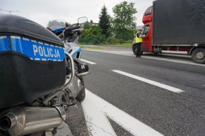 Na pierwszym planie bok kufra z napisem policja na motocyklu, w tle policjant rozmawia z kierowcą ciężarówki.