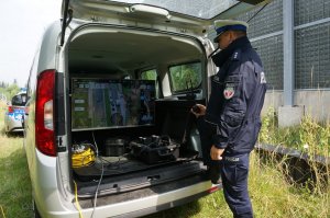 Policjant stoi z tyłu samochodu i obserwuje ekran monitora, na którym widać podgląd z kamery drona.