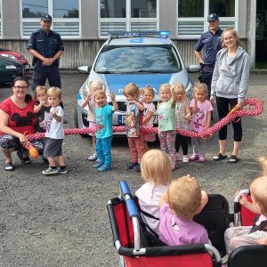 Policjanci stoją przy radiowozie przed budynkiem przedszkola, obok nich i przed nimi dzieci razem z opiekunami.