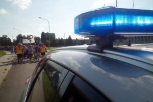 Widok na dach policyjnego radiowozu z włączonym światłem niebieskim, w tle drogą idą pielgrzymi.
