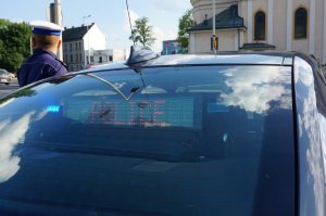 Pod tylną szybą policyjnego radiowozu nieoznakowanego wyświetla się napis POLICJA, obok radiowozu stoi policjant ruchu drogowego