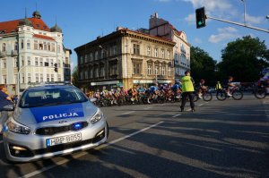 Na skrzyżowaniu stoi radiowóz, w tle peleton kolarski, a przed nimi policjant