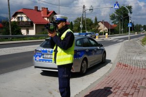 Policjant z ruchu drogowego stoi przy radiowozie i mierzy prędkość pojazdów.