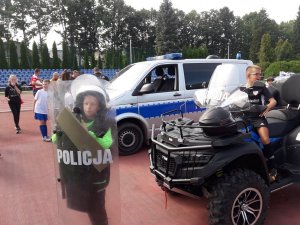 Na stadionie dziecko stoi ubrane z policyjny kombinezon i trzyma tarczę z pleksi, obok siedzi dziecko na policyjnym quadzie.