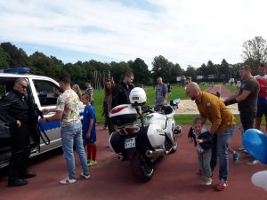 Przy radiowozie policjant rozmawia z dziećmi i rodzicami, obok policyjny motocykl. Zdjęcie wykonane na stadionie.