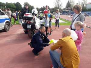 Policjant w mundurze przykuca obok dziecka, przy nim rodzic. Zdjęcie wykonane na stadionie.