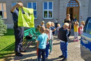 Policjant pokazuje dzieciom kamizelkę odblaskową tuż przy radiowozie.