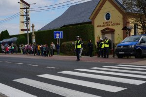 Policjant z drogówki wskazuje tarczą drogową miejsce do zatrzymania pojazdu, na drugim planie przy przystanku stoją dzieci ze szkoły podstawowej.