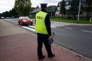 Policjant z ruchu drogowego z ręcznym miernikiem prędkości obserwuje drogę, którą jedzie czerwony samochód osobowy.