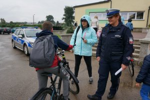 Policjantka ruchu drogowego rozmawia z dzieckiem na rowerze, stoją przed szkołą podstawową.