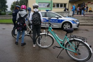 Policjantka ruchu drogowego rozmawia z uczniem, stojącym przy rowerze. Stoją przed szkołą, na drugim planie radiowóz.