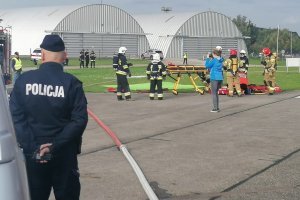 Widok na policjanta stojącego tyłem, na drugom planie trwają ćwiczenia służb na lotnisku.