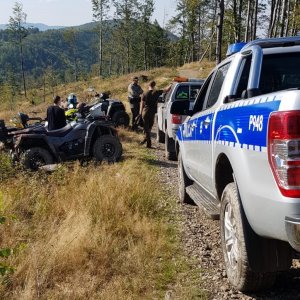 Na pierwszym planie policyjny radiowóz, w tle strażnicy leśny przy pojazdach terenowych i quadach. Zdjęcie wykonane na górskim szlaku.