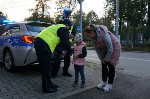 Policjantka ruchu drogowego stoi za radiowozem i zakłada opaskę odblaskową dziecku z matką.