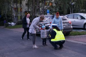 Policjantka kuca przed dzieckiem i z nim rozmawia, obok jego opiekunki. Zdjęcie wykonane na osiedlowej drodze.