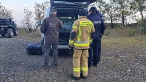 Policjant, strażak i strażnik leśny stoją przy samochodzie technicznym obsługującym drona. Zdjęcie wykonane w lesie.