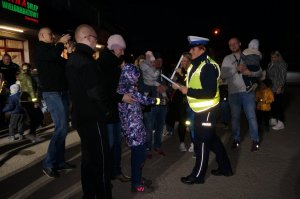 Policjantka z ruchu drogowego zakłada dziecku opaskę odblaskową na rękaw. Obok inni uczestnicy akcji. Zdjęcie wykonane w nocy.
