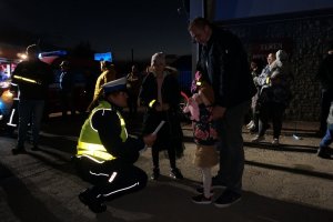 Policjantka z ruchu drogowego kuca przy dziecku i zakłada mu opaskę odblaskową na rękaw. Obok inni uczestnicy akcji. Zdjęcie wykonane w nocy.
